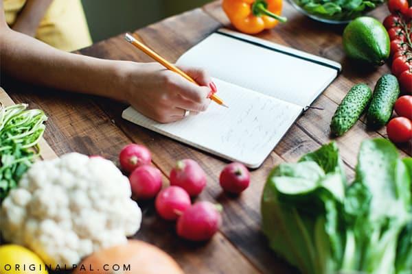 نوشتن میزان کالری و فعالیتهای روزمره در دفتر بر روی میز پر از میوه و سبزیجات